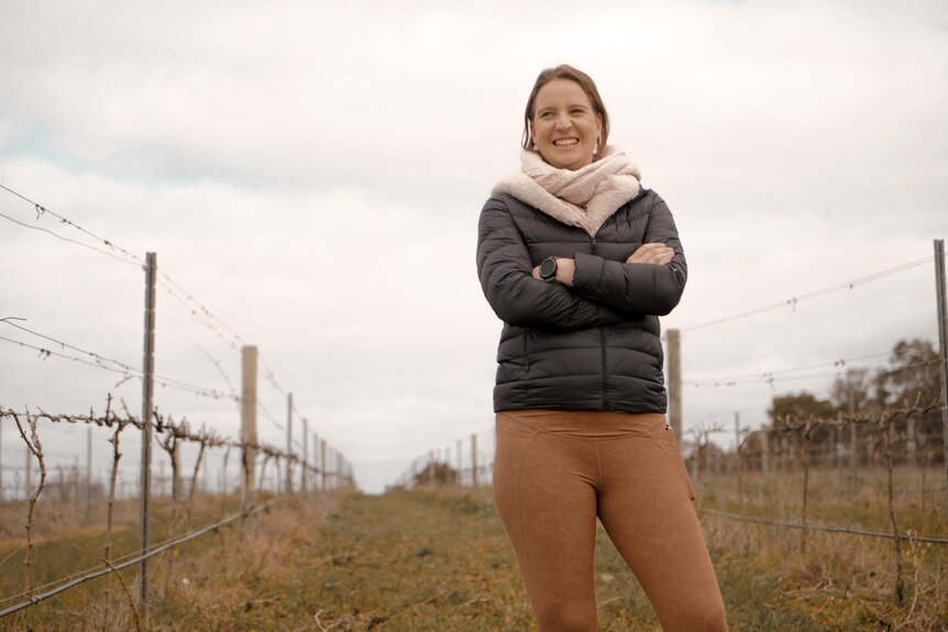 A woman smiles as she crosses her arms while standing in a vineyard.