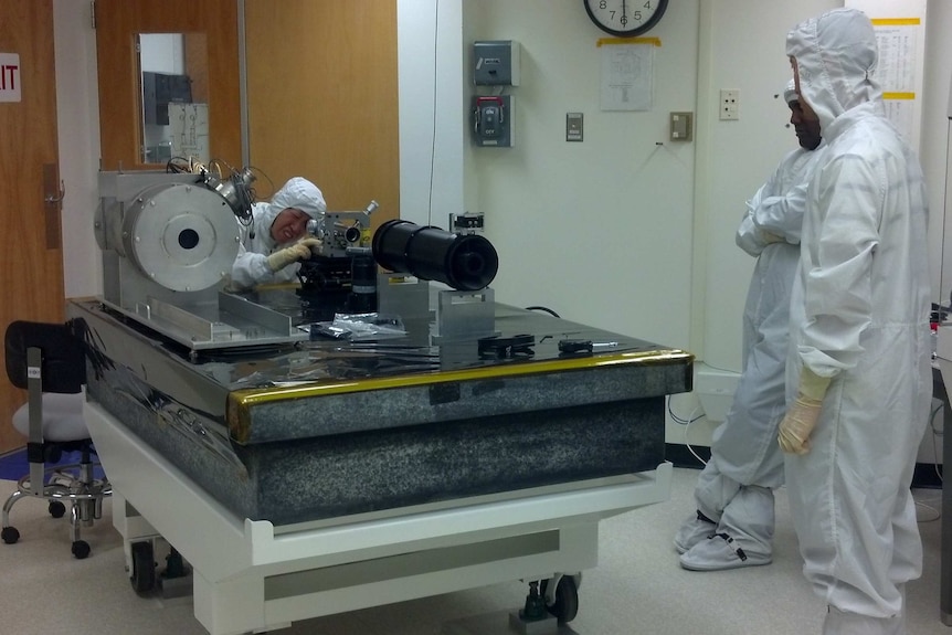 People in white lab suits work on a device on a table