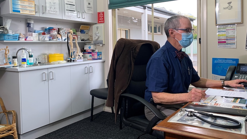 Man wearing face mask at a desk with a stethoscope, looking at a computer. Shelf behind has model of human skeleton, medicines.