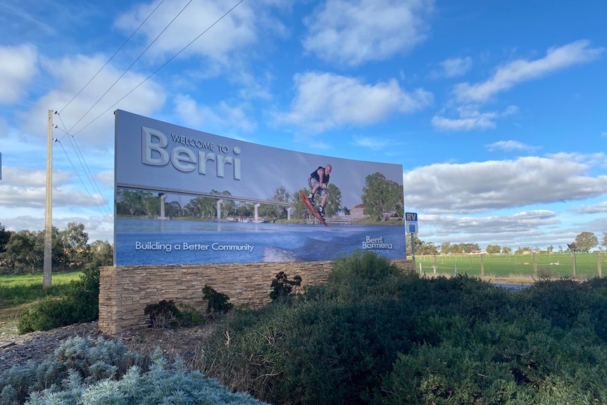 A blue sign reads 'Welcome to Berri', with bushes in front of it and a cloudy sky behind it.