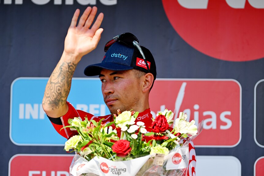 Caleb Ewan holds up his hand and looks to one side standing behind a bunch of flowers