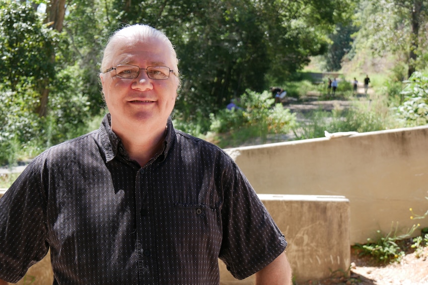 A middle aged man wearing glasses and a back shirt stands near concrete barriers with a damaged road behind him.