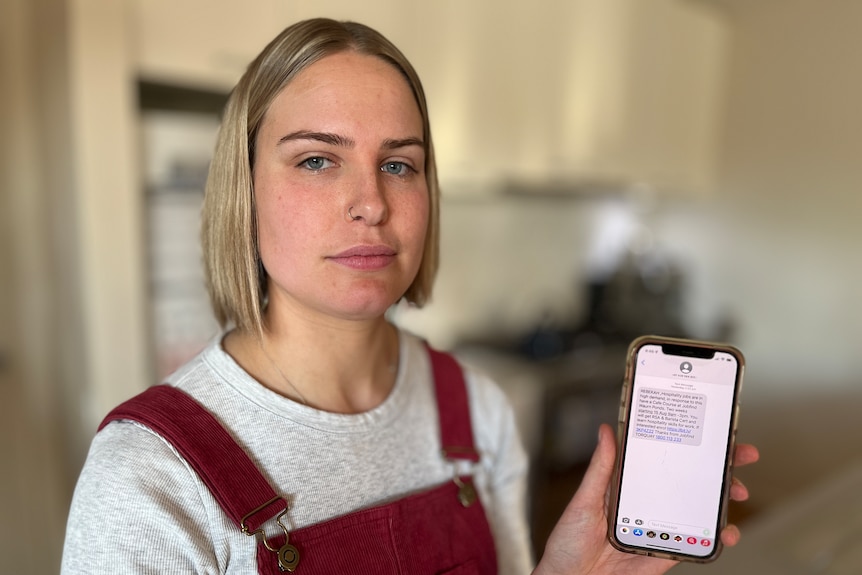 A woman with blonde hair holding up her phone, looking at the camera