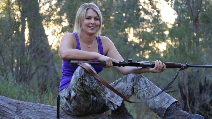 A woman in camo pants and a purple tank top, sitting on a log, holding a gun.
