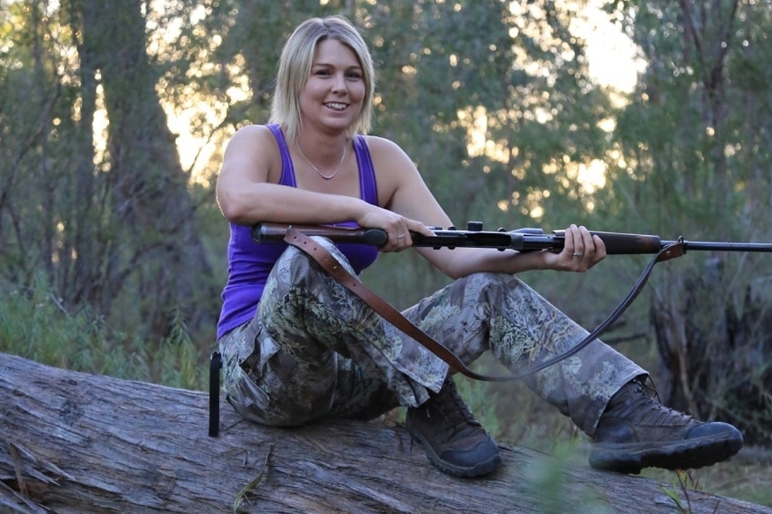 A woman in camo pants and a purple tank top, sitting on a log, holding a gun.