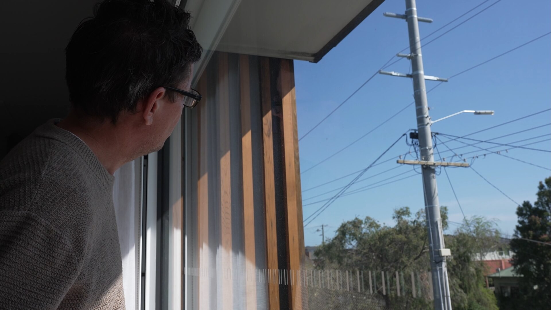 A man in a grey sweater looking out a window to see a large power pole. 