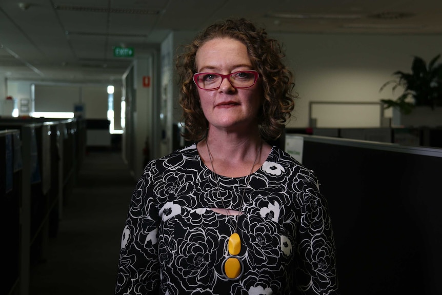 A woman wearing brightly-coloured glasses and a brightly-coloured necklace stands in a dark office building.