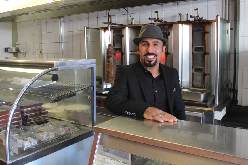 A man smiles at the camera in front of kebabs.