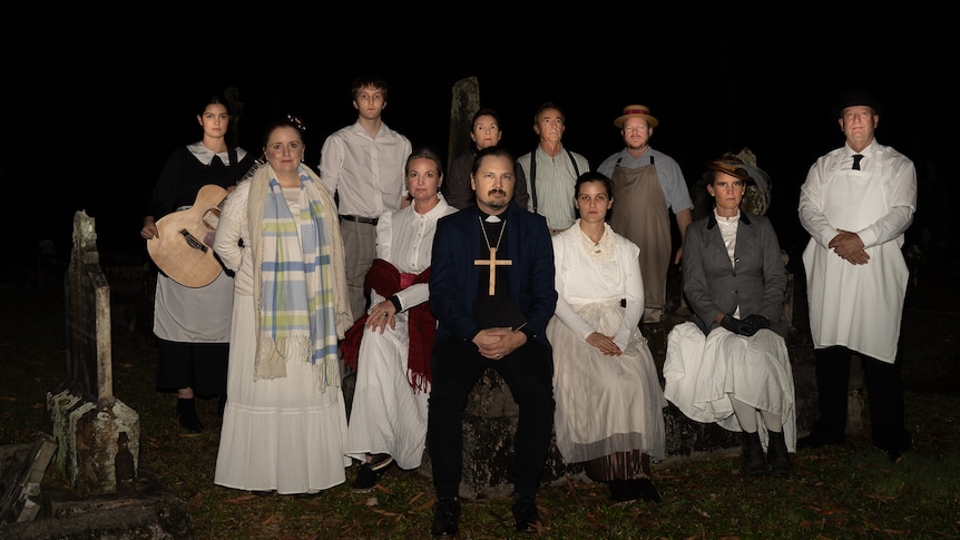 Group of people in costume,. including a priest with a big cross, women dressed in Victorian clothes, background pitch dark. 