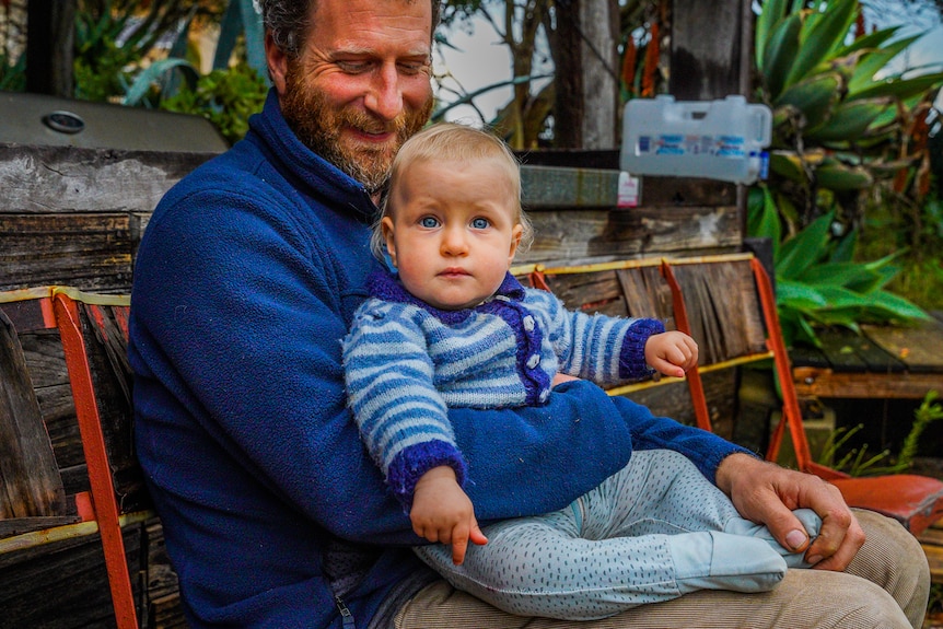 A man in a blue jumper sits outside holding a baby, smiling.