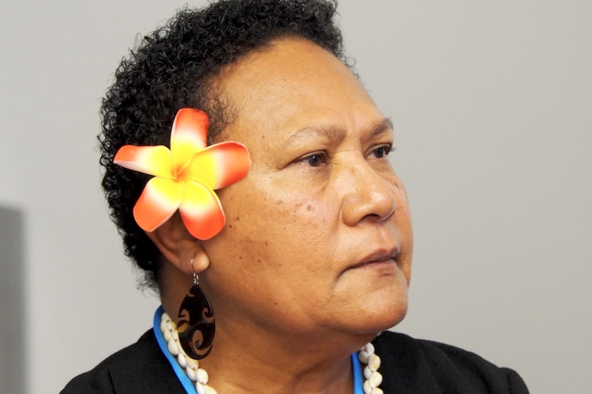 A headshot of a woman of Pacific heritage with short curly dark hair and a large yellow and orange flower behind her ear.