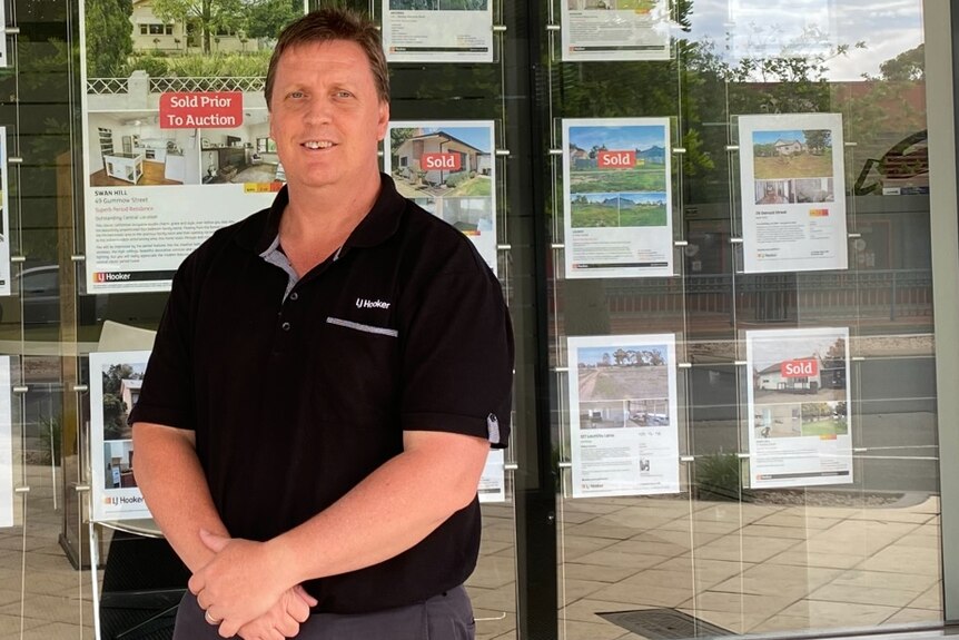 Real estate agent standing out the front of his shop smiling with property ads in the background