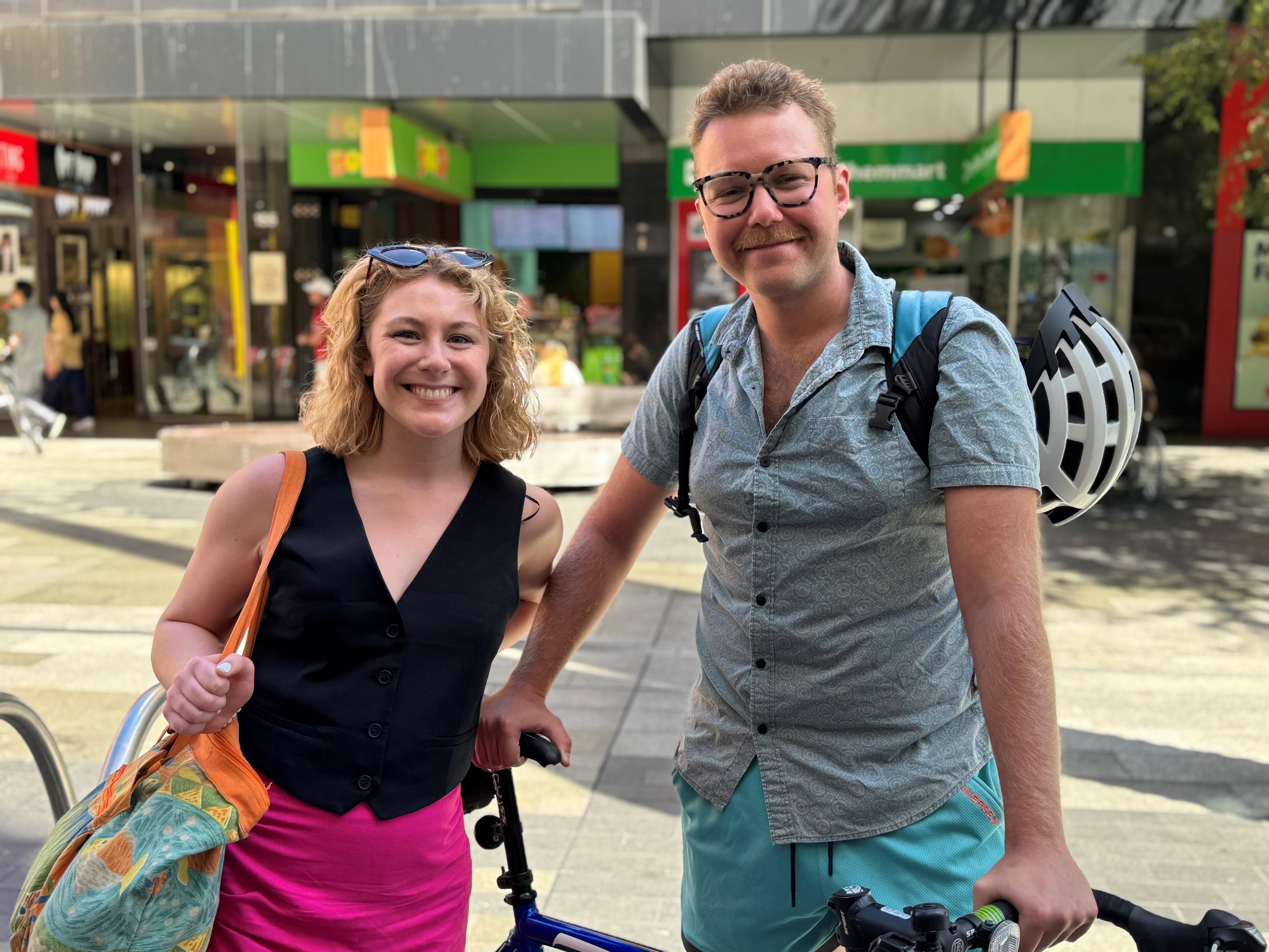 Two people smiling and leaning on a bike in an open-air shopping strip
