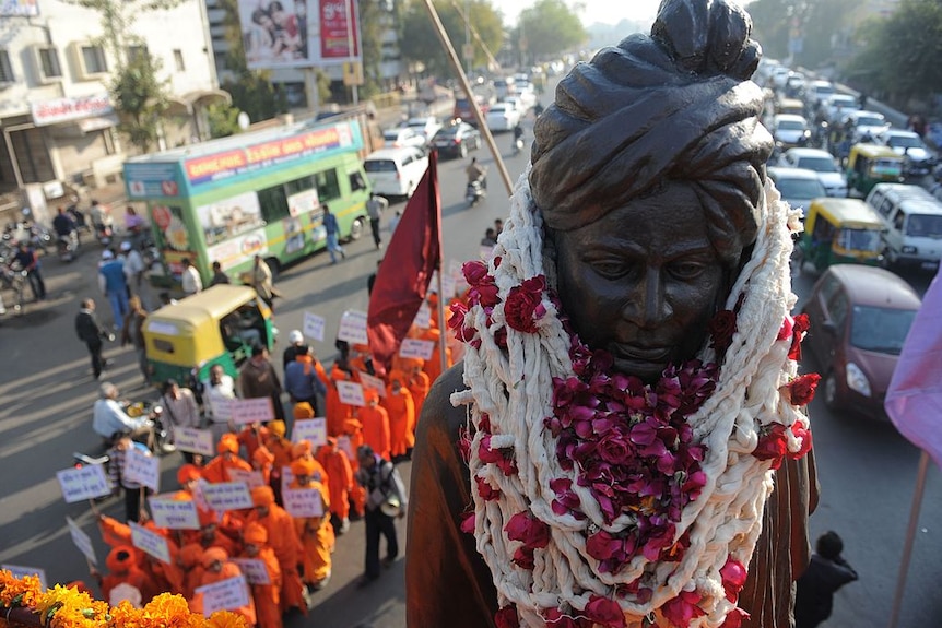Indian school students dress in orange robes like Swami Vivekanand walk down a street alongside his statue.