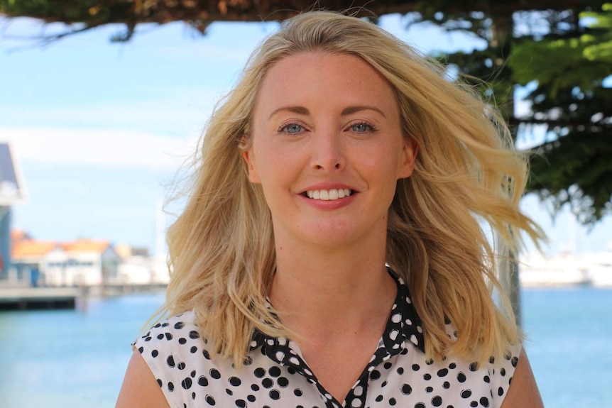 A blonde haired beaming Caitlin Collins smiles at the camera with the ocean in the background.