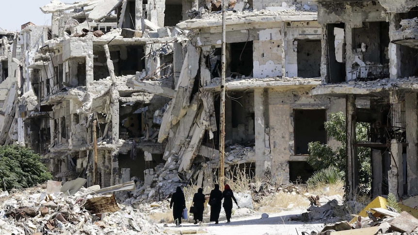 Syrian women walk between destroyed buildings.