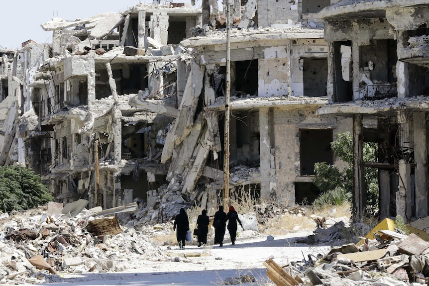 Syrian women walk between destroyed buildings.