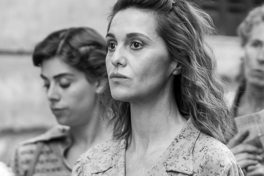 A black and white film still of three women in the street. At the centre, one looks forward with a sense of determination.