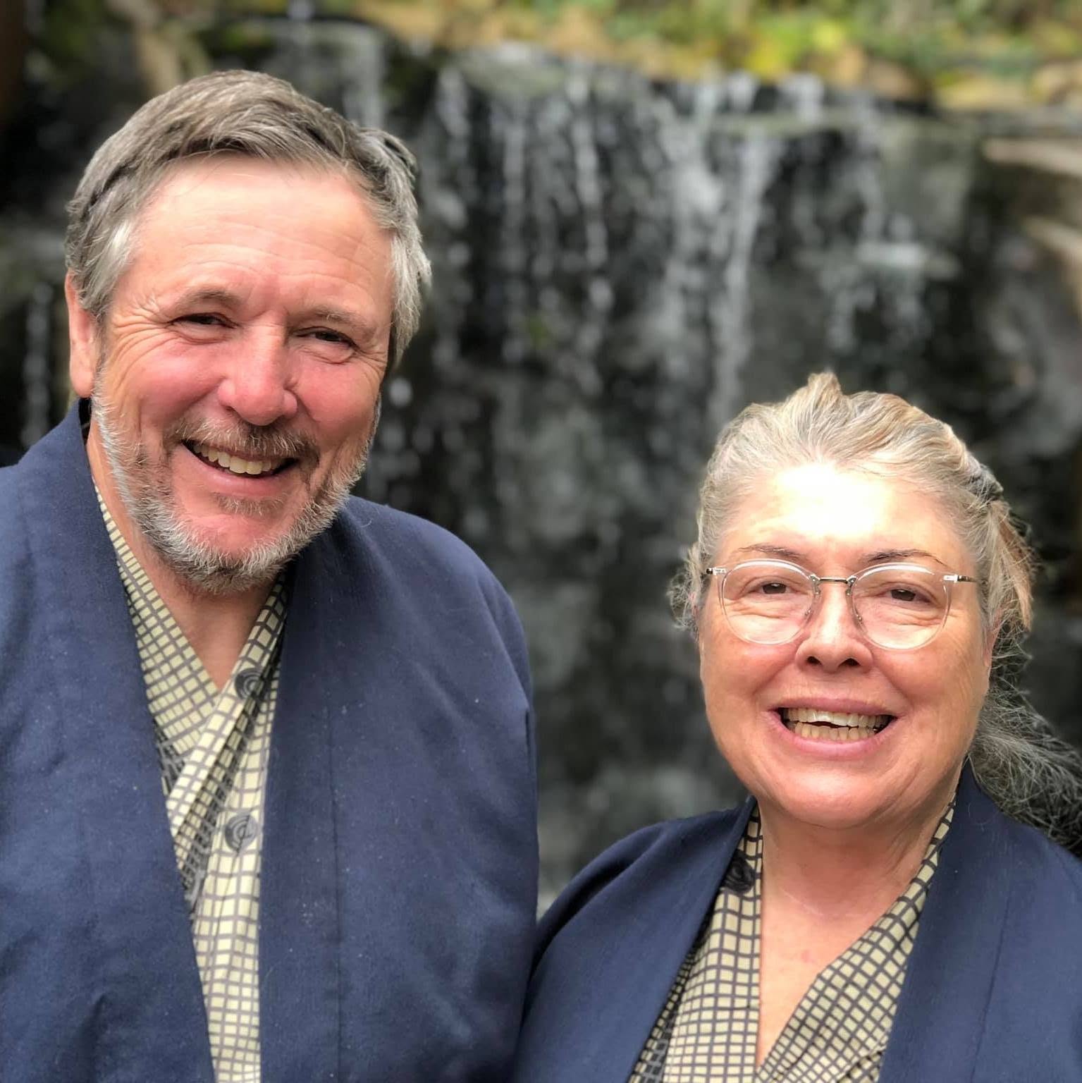 A husband and wife stand in front of a waterfall in Japan and are both wearing traditional Japanese clothing.