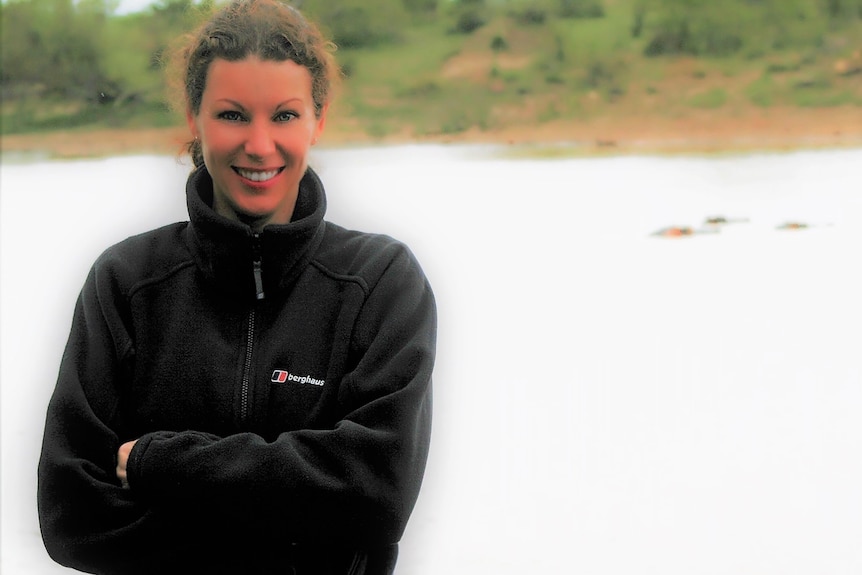 A young women poses smiling with her arms crossed by a body of water.