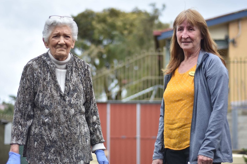 A woman with white hair and purple plastic gloves on her hands stands near a woman with red hair and a yellow shirt