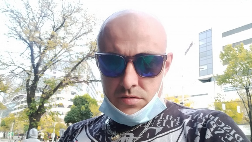A bald man wearing sunglasses and a mask around his chin takes a selfie.