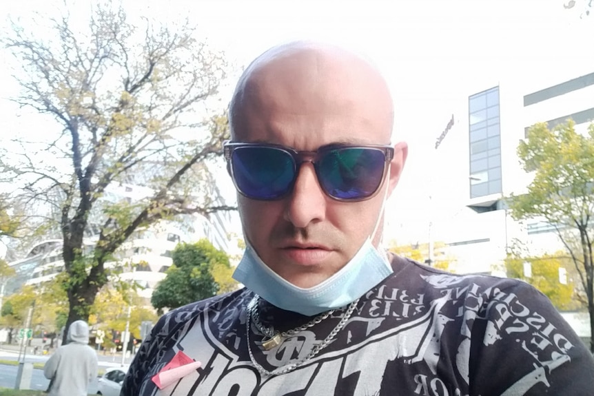 A bald man wearing sunglasses and a mask around his chin takes a selfie.