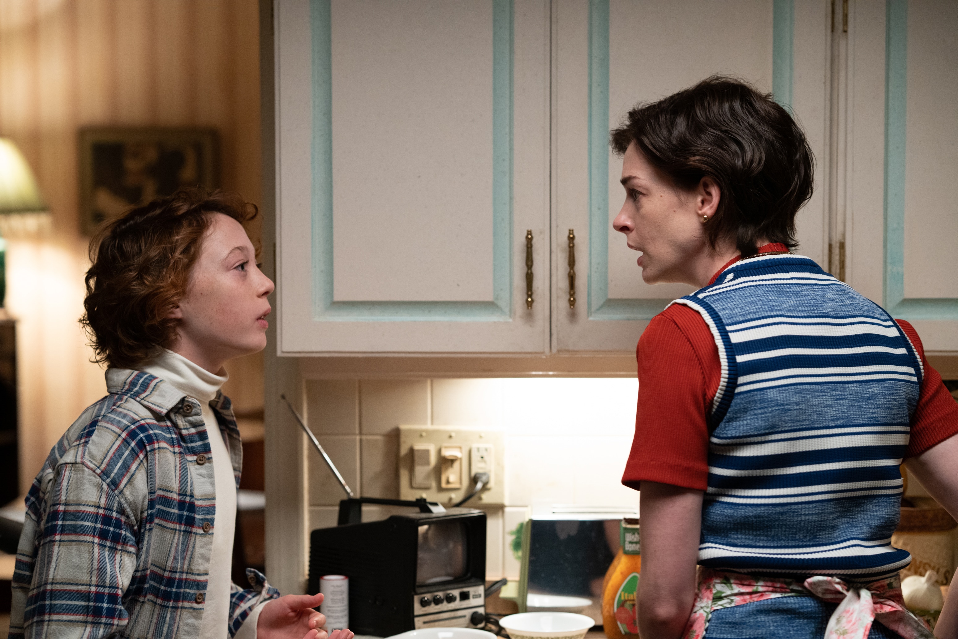 Young white boy with reddish hair wears plaid shirt in kitchen beside his mother; a white woman in red shirt and blue vest.