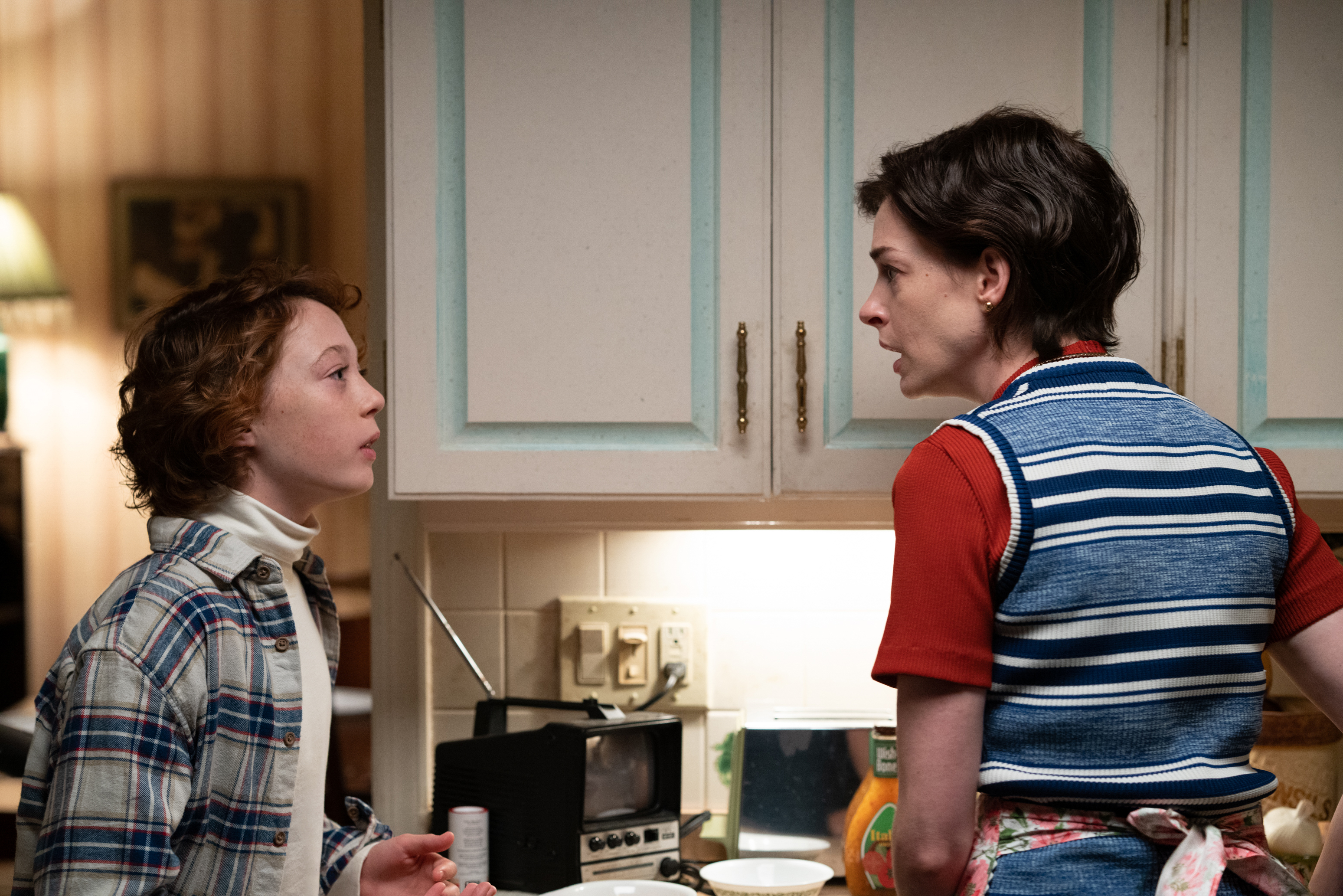 Young white boy with reddish hair wears plaid shirt in kitchen beside his mother; a white woman in red shirt and blue vest.