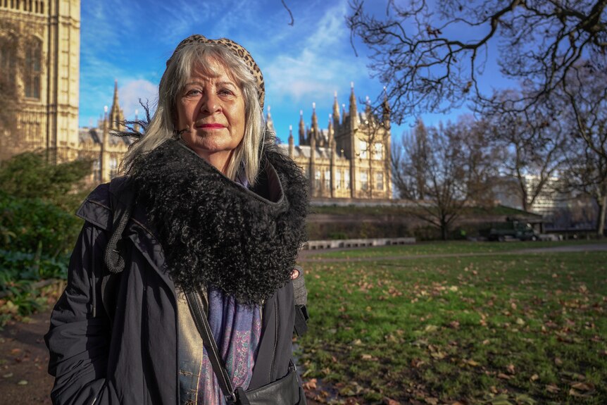 A woman with grey hair and wearing red lipstick and a fur coat stands in a park.