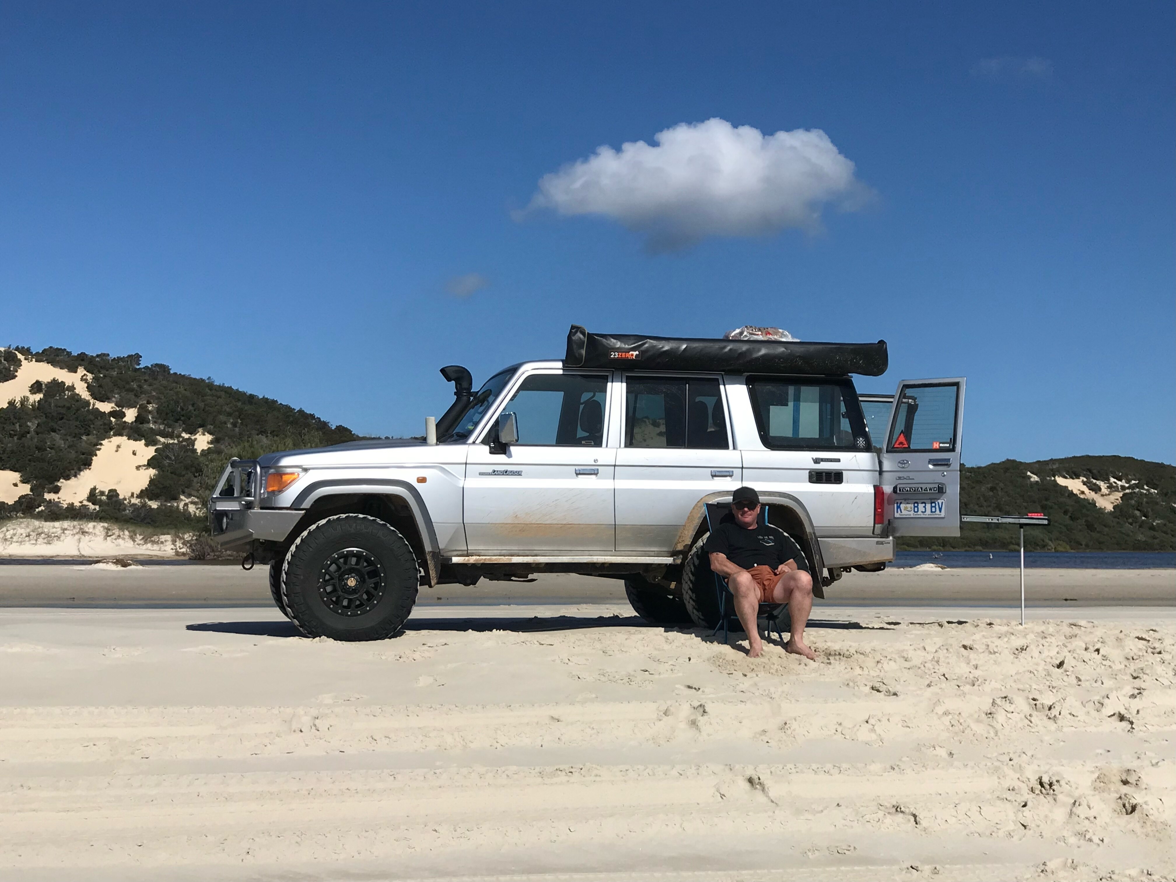 Un hombre se sienta en una silla de campamento frente a un 4WD plateado estacionado en una playa.