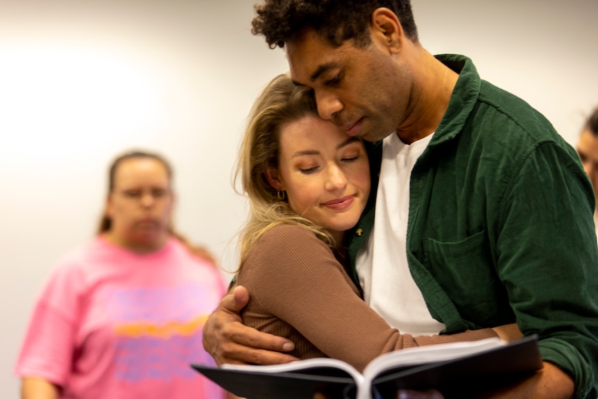 White woman and Indigenous Australian man share a tender embrace in a rehearsal room holding scripts.