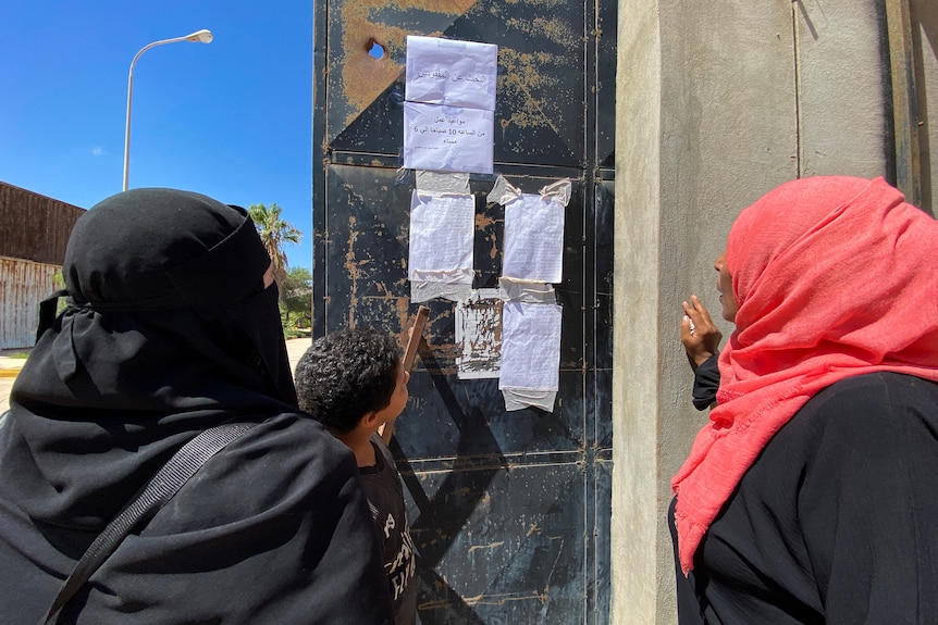 Two women wearing hijab and a young boy look at handwritten paper lists taped to a wall