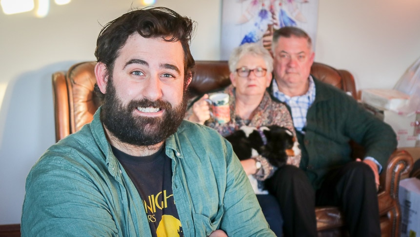 Mike William smiling to the camera, with his parents out of focus in the background on a couch.