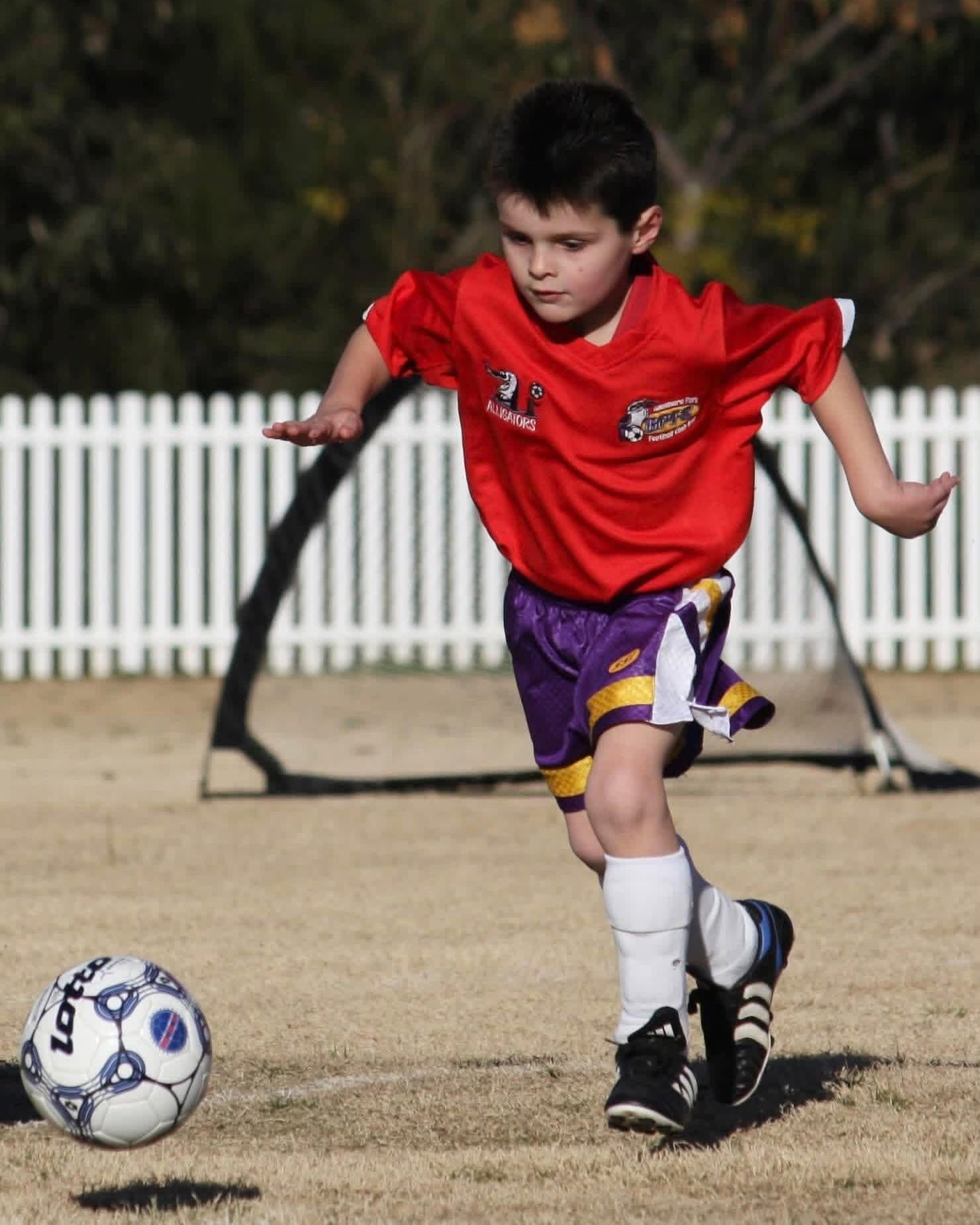 A small boy in a red shirt and purple shorts running while running and kicking a soccer ball in a yard.