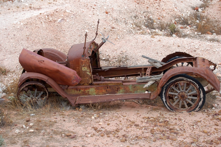 A rusting car with no roof on sand.