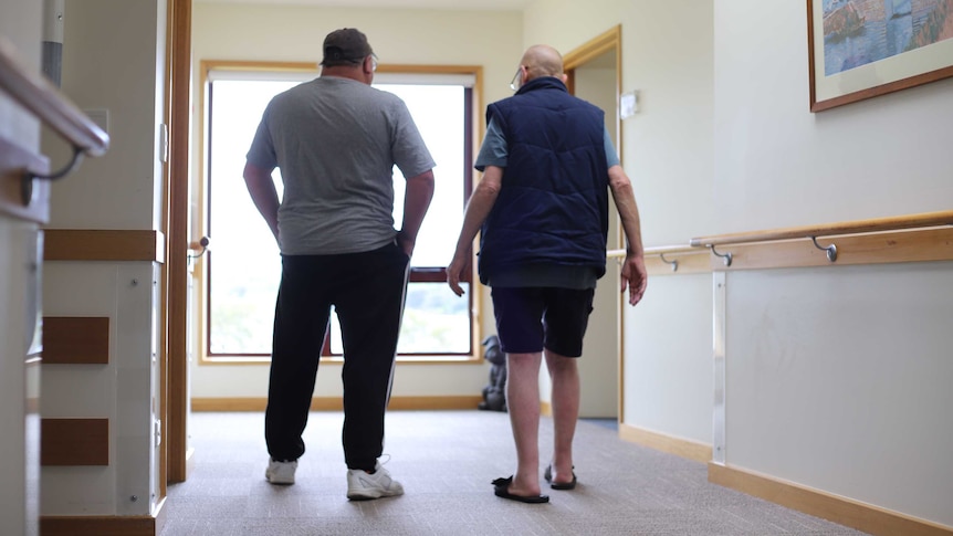 Two men stand side-by-side in a nursing home corridor.
