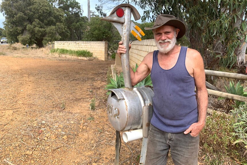 A man wearing a wide brimmed hat stands next to his homemade mailbox - a metal sculpture that resembles an emu.