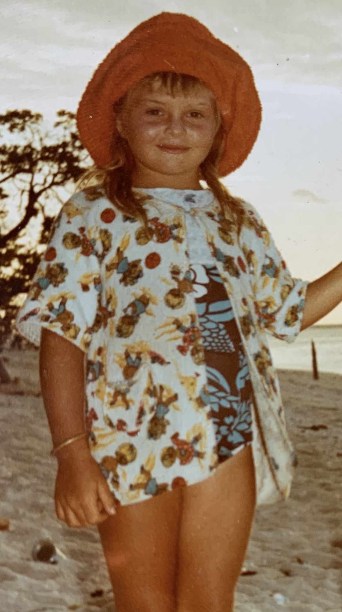 A young blonde girl smiles at the camera wearing swimwear and a floppy red sunhat.
