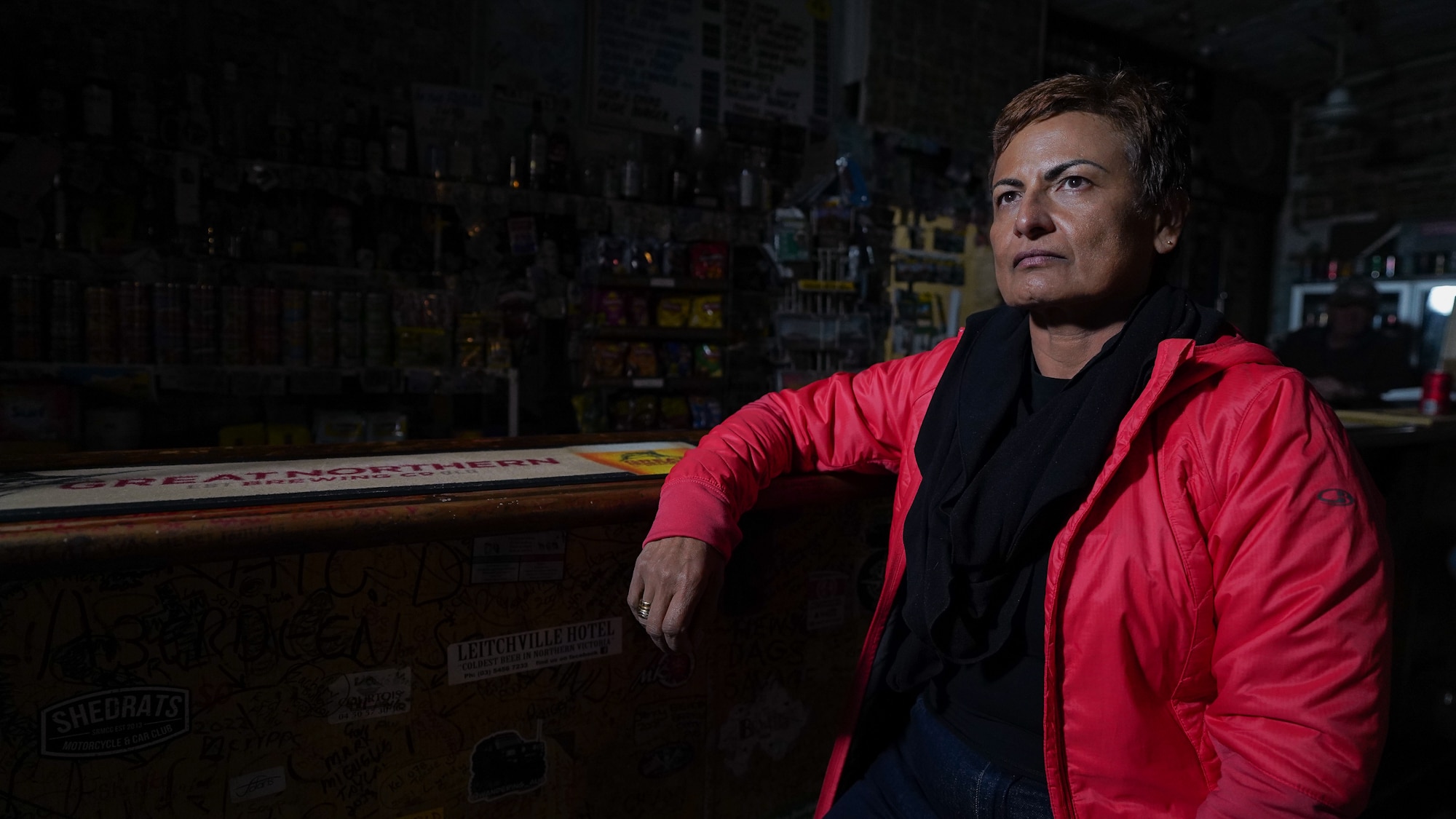 Sitting on a bar stool and leaning on the countertop, a woman in a red jacket wears a tense facial expression.