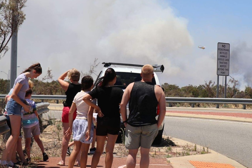 A crowd watching a helicopter hovering over a bushfire.