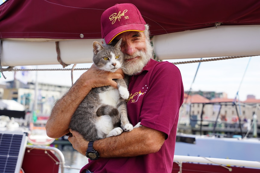 A man with a white beard in a maroon uniform holds up and hugs a cat.