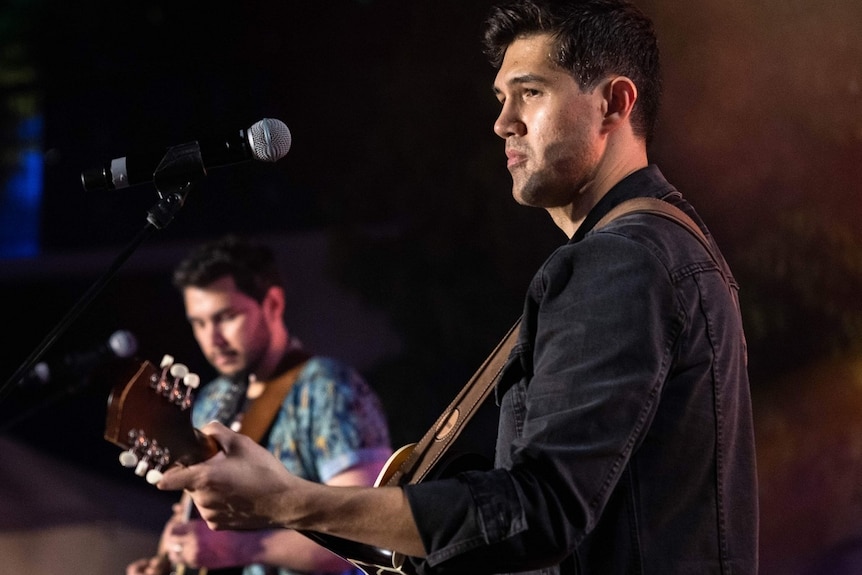 A side on view of Angelo Conway as he plays the guitar on stage.