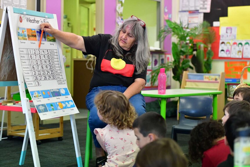A woman in an Aboriginal flag t-shirt points to a calendar on an a-frame as children in a brightly coloured classroom look on.