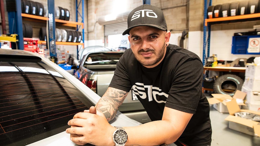 A man leans over the boot of a car in a garage and looks at the camera.