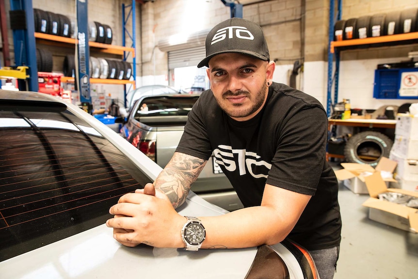 A man leans over the boot of a car in a garage and looks at the camera.