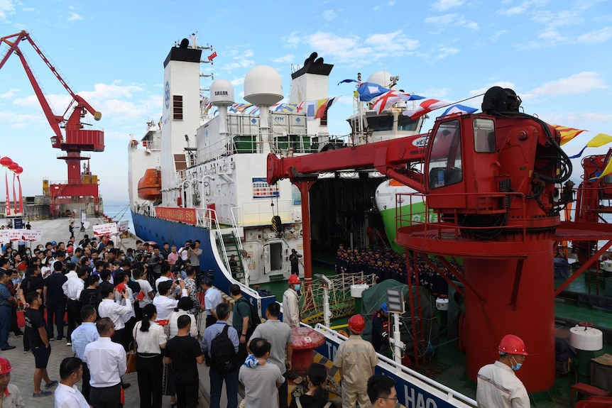 Spectators line a dock to watch submariners disembark a large ship that is carrying their submersible vessel