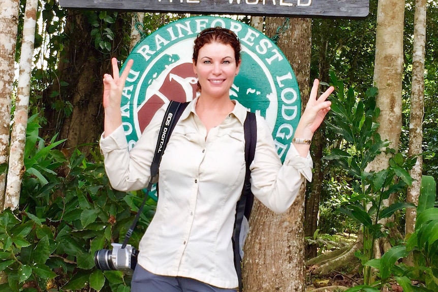 A young woman makes peace signs with her hands as she poses in front of a jungle lodge.