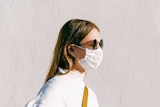 A woman side-on to the camera, wearing sun glasses and a face mask, with a plain wall in the background.