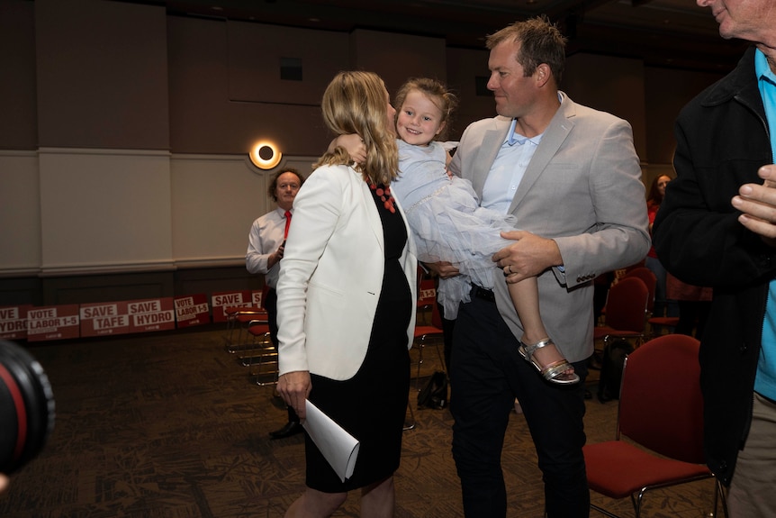 A smiling child held by a man, is kissed by a pregnant woman